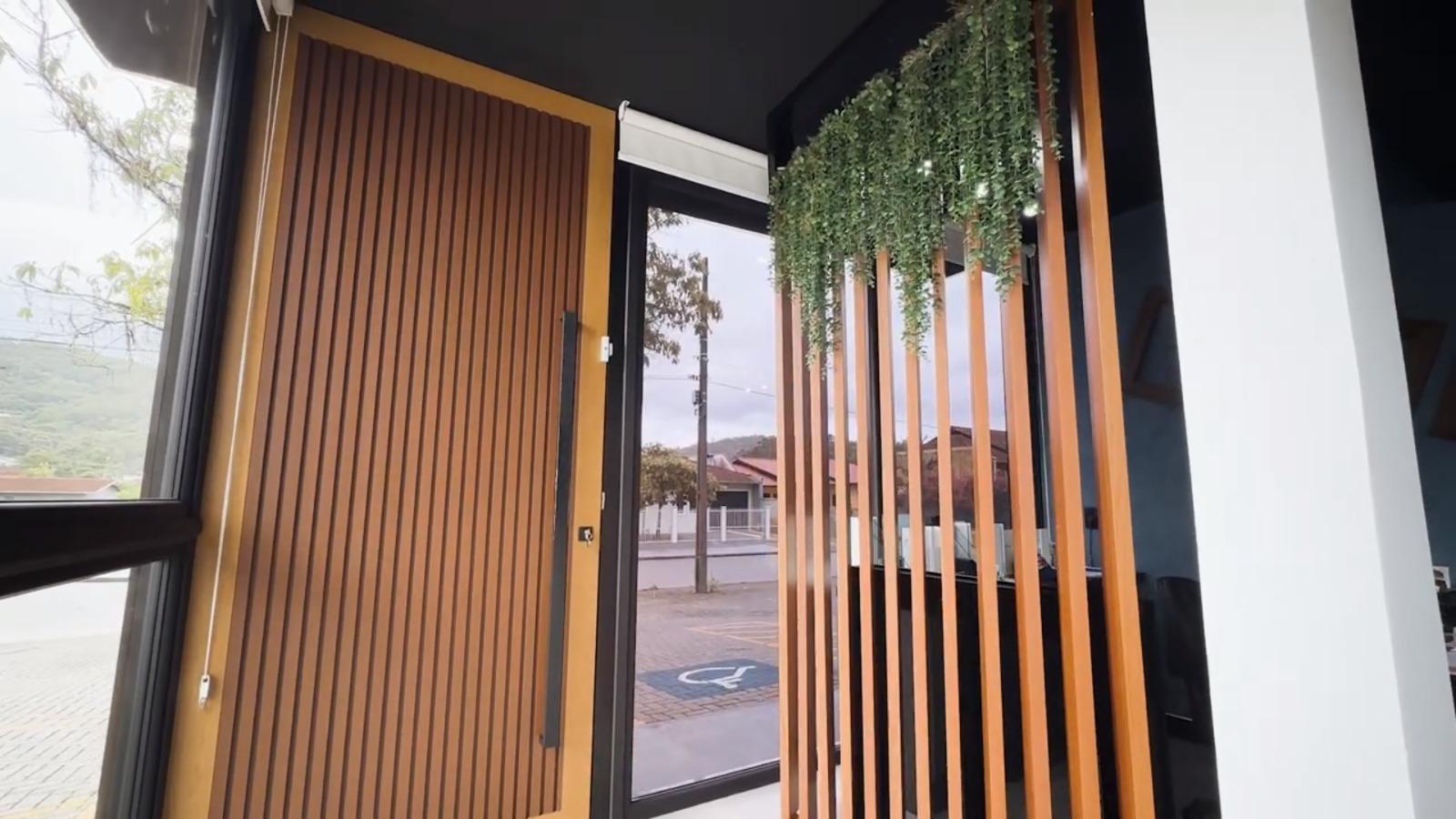 <p> Vista interna de um escritório com decoração de madeira. A imagem mostra uma porta de madeira aberta ao lado de uma janela grande com persiana branca. Há uma divisória de ripas de madeira vertical com plantas artificiais penduradas. Do lado de fora, pode-se ver uma rua com árvores, casas e uma vaga de estacionamento para deficientes. </p>